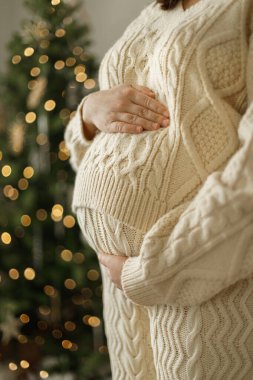 Süslü Noel ağacının arka planında örgü örmüş, göbeğine sarılan mutlu hamile kadın. Tatillerde hamilelik, büyülü neşeli zamanlar.