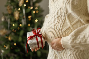 Süslü, örgü kıyafetli, göbeğine sarılan ve elinde Noel hediyesi kutusuyla süslenmiş Noel ağacının arka planına yakın duran hamile bir kadın. Bayramlarda hamilelik, mutlu noeller.