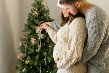 Mutlu hamile noel ailesi, göbeğe sarılıp Noel ağacını birlikte süslüyor. Çiftler arası bağ. Tatillerde hamilelik, büyülü neşeli zamanlar..