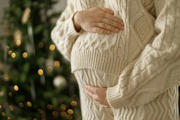 Süslü Noel ağacının arka planında örgü örmüş, göbeğine sarılan mutlu hamile kadın. Tatillerde hamilelik, büyülü neşeli zamanlar.