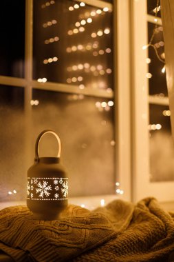 Mutlu Noeller! Şık, ışıl ışıl bir Noel feneri ve altın ışıklar akşam karlı pencerede rahat bir süvetere takılıyor. Atmosferik büyülü sahne. Noel tatili afişi. Metin için boşluk