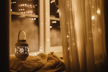 Mutlu Noeller! Şık, ışıl ışıl bir Noel feneri ve altın ışıklar akşam karlı pencerede rahat bir süvetere takılıyor. Atmosferik büyülü sahne. Noel tatili afişi. Metin için boşluk