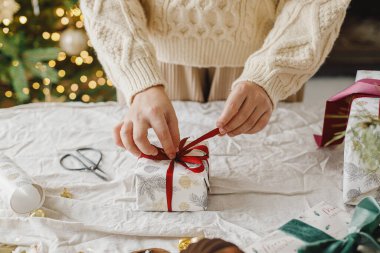 Modern oturma odasındaki dekore edilmiş stil Noel ağacının arka planında keten kumaşla Noel hediyesi paketini hazırlayan eller. Mutlu Noeller ve mutlu Noeller.