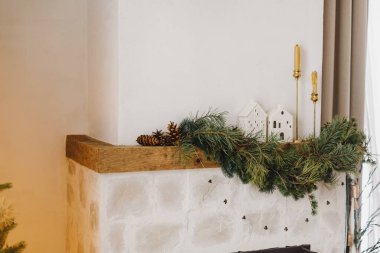Süslü Noel şöminesi şömine rafı köknar dalı, mumlar, seramik evler, çanlar, kurdele. Modern İskandinav odasında şenlikli Xmas kış dekoru. Mutlu Noeller.!