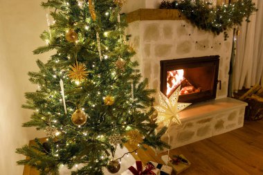 Şık dekore edilmiş yılbaşı ağacı, eski süsler ve şömine rafının modern dekorasyonuyla akşamları çan, kurdele ve çelenk ışıkları. Evde atmosferik Noel arifesi. Mutlu Noeller.!