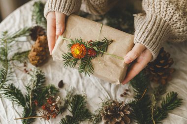 Sıfır Noel konsepti. Tarz sahibi Noel hediyelerini, kır masasında, doğal süslemelerle köknar dalıyla saran eller. Sürdürülebilir hediyeler, kış tatili hediyesi