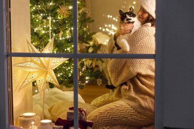 Mutlu Noeller! Mutlu kadın, rahat süveter ve Noel Baba şapkası içinde Noel ışıkları ve hediyelerle pencerede sevimli kedilere sarılıyor. Evde atmosferik Noel arifesi. Tatil büyülü sahnesi