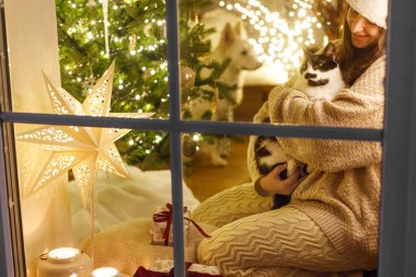 Mutlu Noeller! Mutlu kadın, rahat süveter ve Noel Baba şapkası içinde Noel ışıkları ve hediyelerle pencerede sevimli kedilere sarılıyor. Evde atmosferik Noel arifesi. Tatil büyülü sahnesi