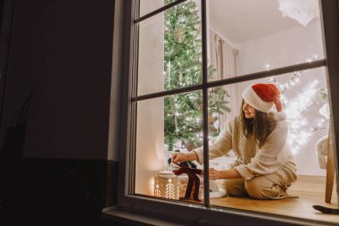 Noel Baba şapkalı güzel bir kadın Noel ağacının altına ışıklarla dolu bir oturma odasının arka planında şık bir Noel hediyesi koyuyor. Evde atmosferik Noel arifesi. Mutlu Noeller.