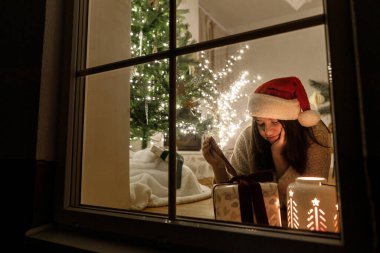 Vitrinde Noel Baba şapkalı mutlu bir kadın şık hediyeler ve arka planda ışıldayan bir fenerle şenlikli oturma odasında ışıkları olan bir Noel ağacı. Evde atmosferik Noel arifesi. Mutlu Noeller.