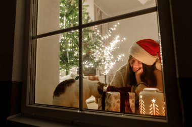 Mutlu Noeller! Noel Baba şapkalı mutlu kadın ve penceresinde şık hediyeli şirin kedi ve ışıklandırmalı yılbaşı ağacının arka planında parlayan fener. Evde atmosferik Noel arifesi.