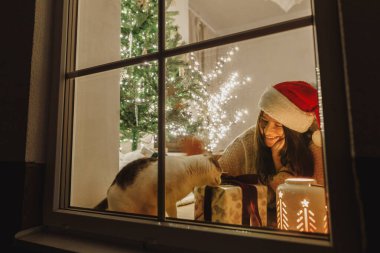 Mutlu Noeller! Noel Baba şapkalı mutlu kadın ve penceresinde şık hediyeli şirin kedi ve ışıklandırmalı yılbaşı ağacının arka planında parlayan fener. Evde atmosferik Noel arifesi.