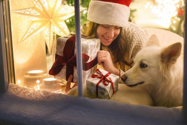 Noel Baba şapkalı, şık bir Noel hediyesiyle oturan mutlu bir kadın ve Noel ağacı ışıklarıyla şenlik odasının içinde karlı pencerede duran sevimli bir köpek. Evde atmosferik Noel arifesi. Mutlu Noeller.!