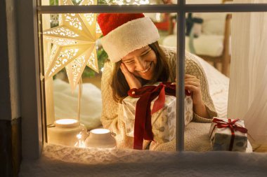 Mutlu Noeller! Rahat süveter ve Noel Baba şapkalı mutlu bir kadın pencerenin önünde şık bir hediyeyle oturuyor. Arka planda ışıklı bir festival odası. Evde atmosferik Noel arifesi. Tatil büyülü sahnesi