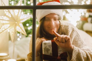 Mutlu Noeller! Noel Baba şapkalı mutlu bir kadın penceresinde parmaklarıyla kalp çiziyor ve arka planda şen şakrak bir hediye var. Evde atmosferik Noel arifesi. Tatil büyülü sahnesi