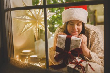 Mutlu Noeller! Noel Baba şapkalı mutlu kadın şık bir hediyeyle pencerede oturuyor ve arka planda şenlik ışıklarının hayalini kuruyor. Evde atmosferik Noel arifesi. Tatil büyülü sahnesi