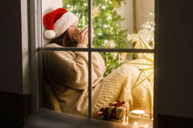 Mutlu Noeller! Noel Baba şapkalı ve rahat kazaklı mutlu kadın, arka planda, Noel ağacı ışıklarının altında sıcak kakao içiyor. Evde atmosferik Noel arifesi. Tatil büyülü sahnesi