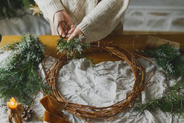 Noel kırsal çelengi yapıyorum. Ebediyete kadar uzanan dalları tutan eller, kozalaklar, sicim, makas ve mum ile kırsal masada çelenk yapıyor atmosferik festival odasında. Kış tatili hazırlıkları