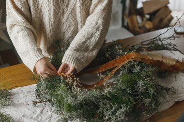 Noel kırsal çelengi yapıyorum. Eller kurdele tutuyor, atmosferik festival odasında kırsal masada çelenk süslüyor. Kış tatili hazırlıkları