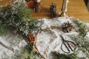 Şık, sade bir Noel çelengi yapıyorum. Evergreen dalları, çam kozalakları, dal, sicim, makas atmosferik festival odasında kırsal masada. Kış tatili hazırlıkları, ayrıntılar