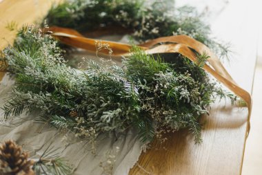 Şık, sade bir Noel çelengi yapıyorum. Köknar ve yeşil dallı çelenk, kurdele, sicim, makas atmosferik festival odasındaki kırsal masada. Kış tatili hazırlıkları