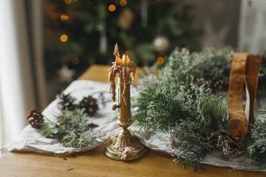 Şık, sade bir Noel çelengi yapıyorum. Yeşil dallı çelenk, kurdele, makas ve mum atmosferik noel ağacının arka planında kırsal masada. Kış tatili hazırlıkları