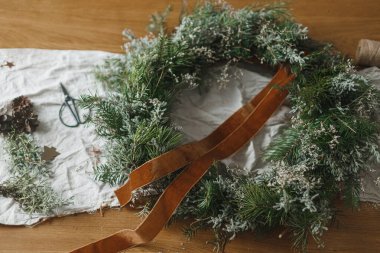 Şık, sade bir Noel çelengi yapıyorum. Köknar ve yeşil dallı çelenk, kurdele, sicim, makas atmosferik festival odasındaki kırsal masada. Kış tatili hazırlıkları