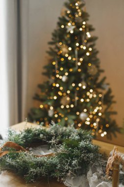 Şık, sade bir Noel çelengi yapıyorum. Yeşil dallı çelenk, kurdele, makas kırsal masada atmosferik noel ağacının arka planında. Kış tatili hazırlıkları