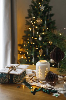 Mutlu Noeller ve mutlu Noeller! Şık bir Noel hediyesi, parlayan bir fener, altın Noel ağacı ışıklarına karşı masanın üstünde modern kağıt süsler. Atmosferik Noel arifesi, büyülü tatil zamanı.