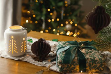Şık bir Noel hediyesi, parlayan fener ve modern kağıt süslemeler arkadaki rustik masa üzerinde Noel ağacı ışıkları bokeh. Mutlu Noeller! Atmosferik Noel zamanı