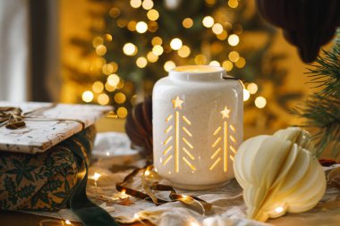 Mutlu Noeller ve mutlu Noeller! Şık bir Noel hediyesi, parlayan bir fener, altın Noel ağacı ışıklarına karşı masanın üstünde modern kağıt süsler. Atmosferik Noel arifesi, büyülü tatil zamanı.