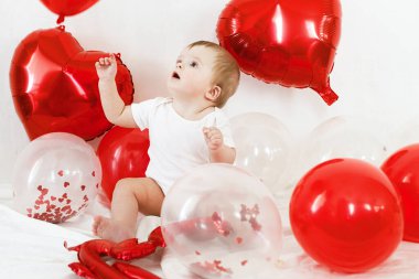 Sevgililer günün kutlu olsun. Kırmızı kalp şeklinde balonların arasında oturan ve gülümseyen sevimli bir bebek. Sevgililer Günü dekorasyonunun arka planında mutlu şirin bir bebek.