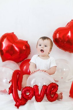 Sevgililer günün kutlu olsun. Kırmızı kalp şeklinde balonların arasında oturan ve gülümseyen sevimli bir bebek. Sevgililer Günü dekorasyonunun arka planında mutlu şirin bir bebek.