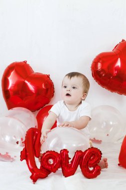 Sevgililer günün kutlu olsun. Kırmızı kalp şeklinde balonların arasında oturan ve gülümseyen sevimli bir bebek. Sevgililer Günü dekorasyonunun arka planında mutlu şirin bir bebek.