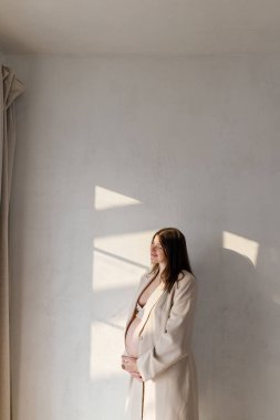 Bej takım elbiseli güzel hamile bir kadın kucağında bebek şişliği ve yumuşak ışıkta duvara karşı poz veriyor. Doğurganlık, sağlık ve yeni yaşam konsepti