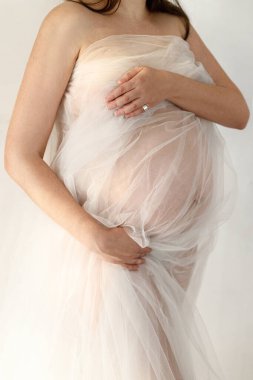 Yumuşak tüller içinde şık hamile bir kadın bebek şişliğine sarılıyor ve arka planda gri duvarda poz veriyor. Doğurganlık, sağlık ve yeni yaşam konsepti