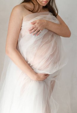 Yumuşak tüller içinde şık hamile bir kadın bebek şişliğine sarılıyor ve arka planda gri duvarda poz veriyor. Doğurganlık, sağlık ve yeni yaşam konsepti