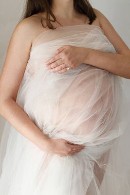 Yumuşak tüller içinde şık hamile bir kadın bebek şişliğine sarılıyor ve arka planda gri duvarda poz veriyor. Doğurganlık, sağlık ve yeni yaşam konsepti