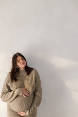 Samimi süveter giyen mutlu hamile kadın, bebek şişliğine sarılıyor ve gri duvarın arka planında duruyor, mesaj için yer var. Kaliteli hamile kadın ve anne. Doğurganlık ve yeni yaşam konsepti