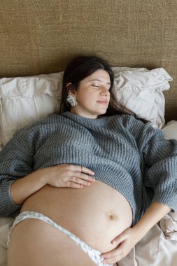 Sabahın köründe yatakta uzanırken kucağında bebek şişliği olan rahat kazaklı hamile bir kadın. Doğurganlık, sağlık ve yeni yaşam konsepti