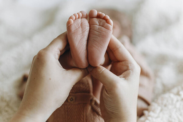Parent hands holding newborn baby feet on cozy blanket background. Love and family concept. Mother holding sweet little infant legs, close up. Adoption and maternity. Child health and care