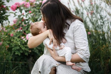 Anne bahçede çiçeklerin arasında sevimli yeni doğmuş bebeği öpüyor ve sarılıyor. Kırsal alanda sevgi, ilgi ve aile. Anneler Günün kutlu olsun! Evlat edinme ve annelik