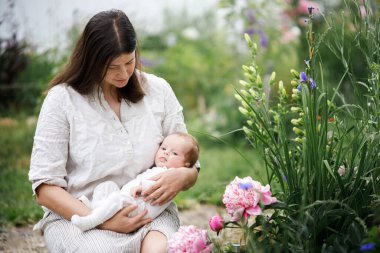 Anneler Günün kutlu olsun! Anne bahçede çiçeklerin arasında sevimli yeni doğmuş bebeğe sarılıyor. Kırsal alanda sevgi, ilgi ve aile. Evlat edinme ve annelik
