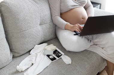 Elinde bebek tamponu olan hamile bir kadın kucağında dizüstü bilgisayarla kanepeye oturmuş bebek kıyafetleri, ultrason fotoğrafı ve oturma odasında kredi kartıyla oturuyor. Hamilelik alışverişi ve hazırlık.
