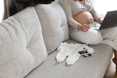 Hamilelik alışverişi ve hazırlık. Bebek karnında bebek kartıyla kucağında dizüstü bilgisayar olan hamile bir kadın bebek elbiseleriyle kanepede otururken, oturma odasında ultrason fotoğrafı....