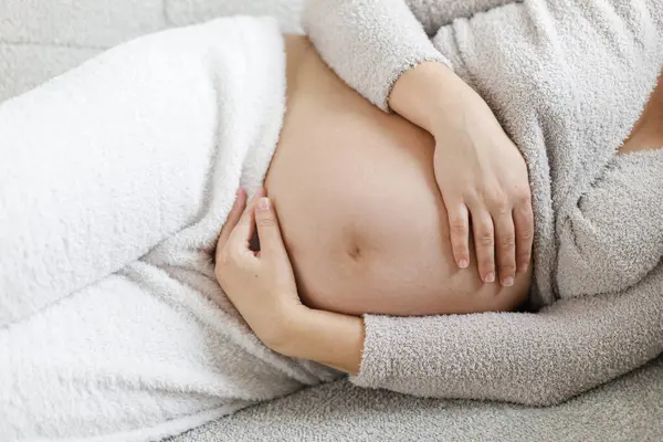 Rahat giysiler içinde hamile bir kadın modern oturma odasındaki gri koltukta bebek şişliğine sarılıp dinleniyor. Doğurganlık ve annelik. Anneler Günün kutlu olsun. Aşk ve bakım konsepti