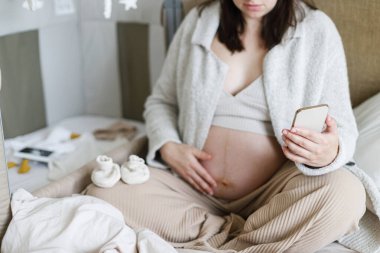 Hamile bir kadın telefon tutuyor ve bebek beşiğine cep telefonuyla sarılıyor. Ultrason fotoğrafı, giysi ve oyuncakla. Hamilelik. Bebek odası hazırlığı ve online alışveriş