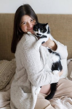 Hamile bir kadın bebek şişliğinde sevimli kediye sarılıyor ve yastıklı rahat bir yatakta oturuyor, kapat. Hayvan ve Hamilelik. Doğurganlık. Kedi arkadaşla bebek hazırlığı