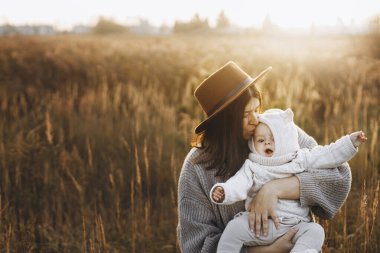Mutlu anne, güz tarlasında, yumuşak akşam ışığında tatlı oğlunu kucaklıyor ve öpüyor. Ebeveynlik. Anne ve küçük çocuk rahat kıyafetler içinde sonbahar parkında kucaklaşıyorlar, atmosferik anları seviyorlar.