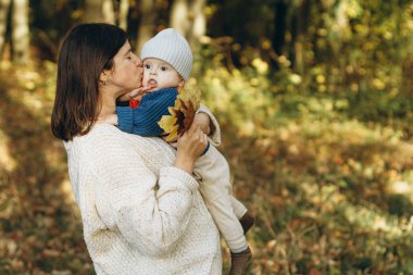 Mutlu anne, sonbahar ormanlarında sevimli oğlunu kucaklıyor ve öpüyor. Annelik. Güz parkında rahat süveter giyen sevimli çocuklu şık bir kadın.